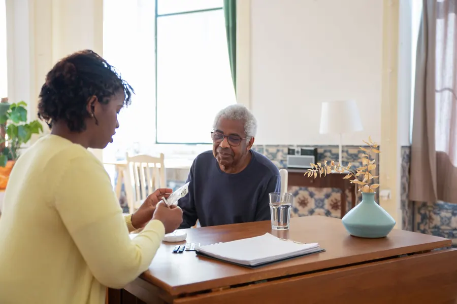 A caregiver sorts a weekly pill organizer at a dining table while an older man looks on
