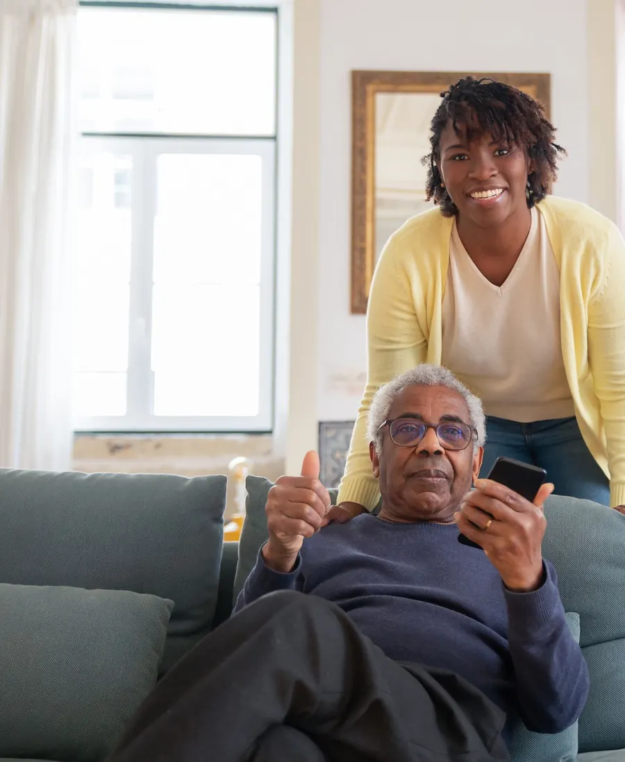 A smiling caregiver in a yellow cardigan leans over the sofa where an older man relaxes in a bright living room