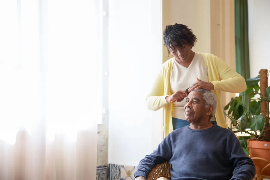 A caregiver gently combs an older man's hair beside a sunlit window