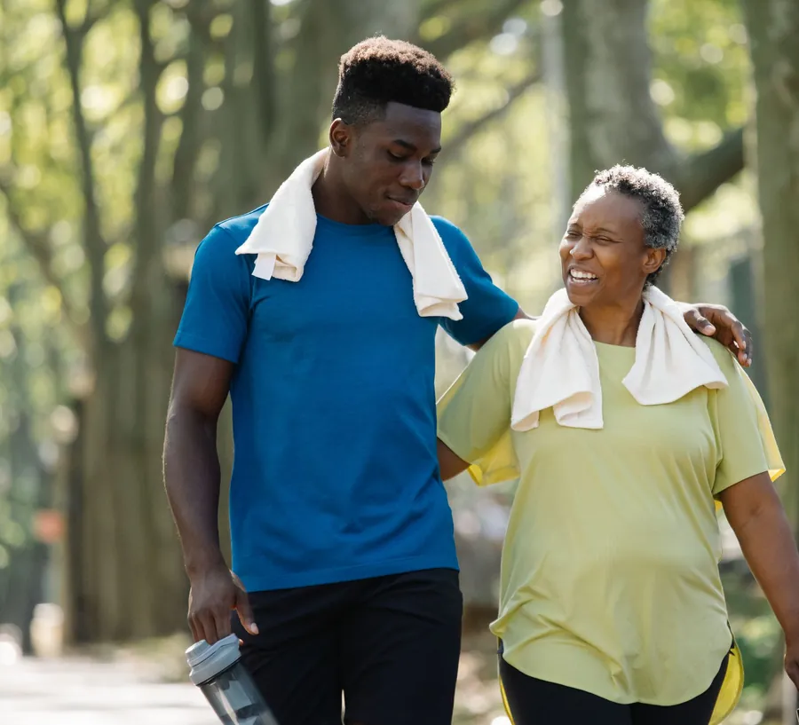 An older woman and a young man walk arm in arm, laughing, along a leafy park path